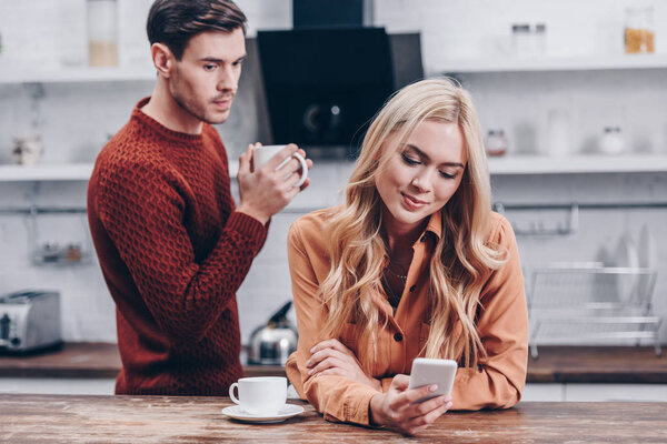 ревнивый молодой человек смотрит на улыбающуюся девушку с помощью смартфона на кухне, концепция недоверия
