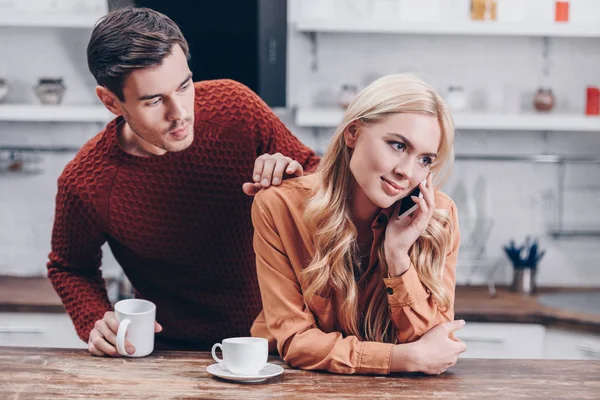 jealous young man looking at smiling wife talking by smartphone in kitchen, mistrust concept