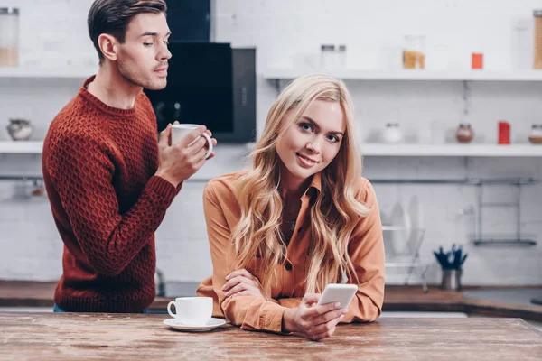 Jealous Young Man Looking Wife Using Smartphone Smiling Camera Kitchen — Stock Photo, Image