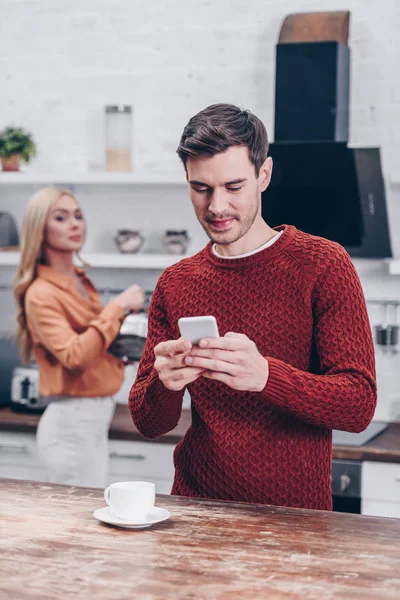嫉妒的女人看着微笑的丈夫在厨房使用智能手机 不信任的概念 — 图库照片