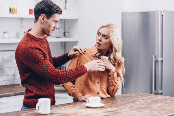 ревнивый молодой человек смотрит на испуганную девушку с помощью смартфона на кухне
