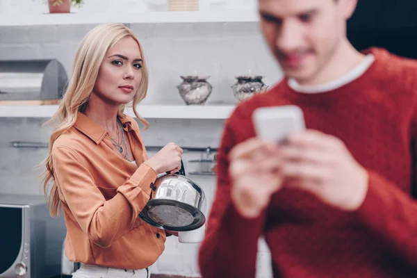 有选择的焦点嫉妒的年轻女子与水壶看丈夫使用智能手机在厨房 — 图库照片