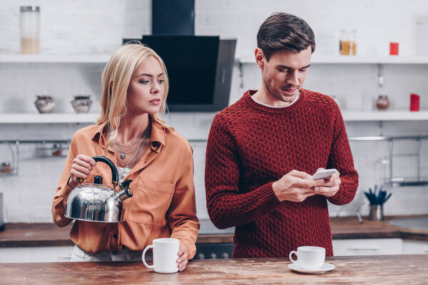 jealous young woman with kettle looking at husband with smartphone in kitchen 