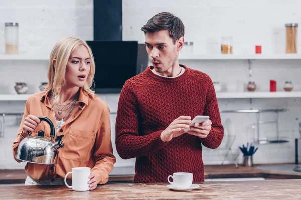 Jealous Young Woman Holding Kettle Looking Husband Smartphone Kitchen — Stock Photo, Image
