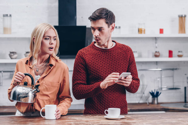 jealous young woman holding kettle and looking at husband with smartphone in kitchen 