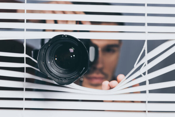 close-up view of man holding camera and peeking through blinds, mistrust concept