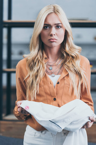 расстроенная молодая женщина держит белую мужскую рубашку со следами помады и смотрит в камеру
 