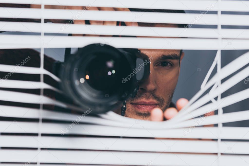 young man holding camera and looking at camera through blinds, mistrust concept