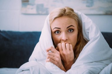scared woman covered in blanket biting hand and looking at camera in bed  clipart