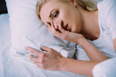 yatakta yatarken, yüz el ile kapsayan ve smartphone kullanarak kadın
