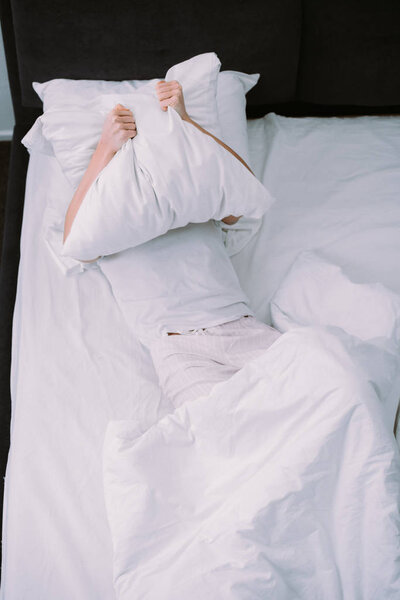 depressed woman covering face with pillow while lying in bed at home