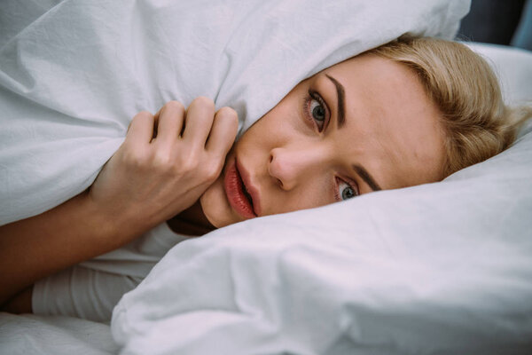 scared woman covered in blanket looking at camera in bed at home