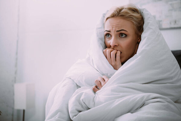 selective focus of scared woman covered in blanket biting hand in bed 