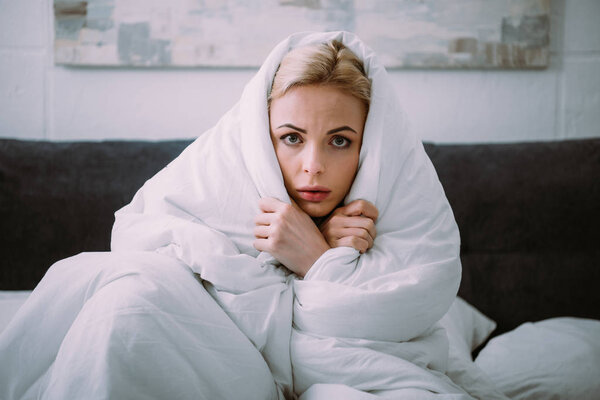 frightened woman covered in blanket looking at camera in bed 