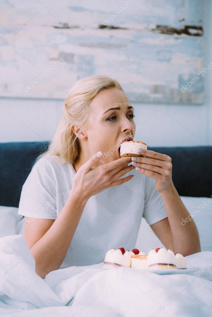 Sad woman in pajamas eating sweet cake in bed alone