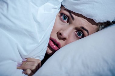 scared woman hiding behind blanket and looking at camera in bedroom at home clipart