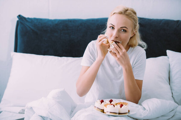 woman looking at camera while crying and eating sweet cake in bed alone