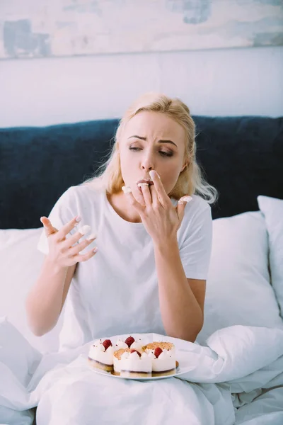 一人ぼっちでベッドでケーキを食べながら指を舐めているパジャマで悲しい女 — ストック写真