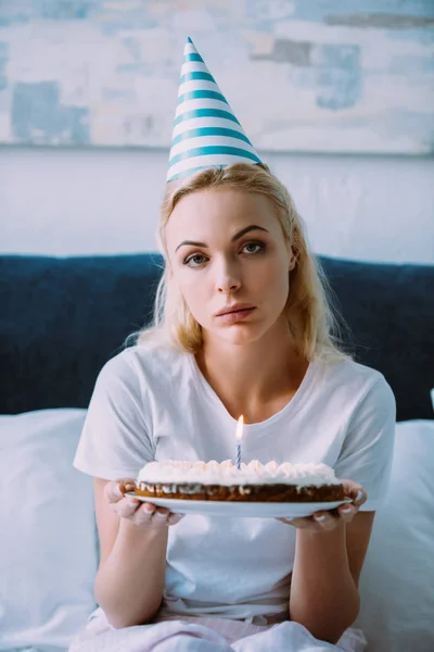 沮丧的女人拿着蛋糕 看着相机 而庆祝生日独自在床上 — 图库照片
