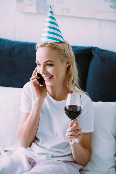 微笑的妇女在党的帽子庆祝生日 拿着一杯葡萄酒 在智能手机上交谈在家里的床上 — 图库照片