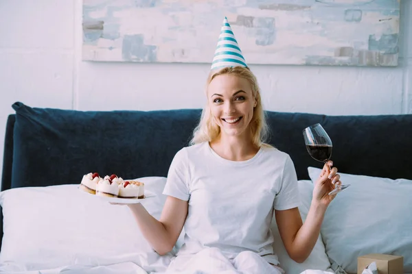 一人ぼっちでベッドで誕生日を祝っている間赤ワインとケーキのガラスを保持パーティー ハットで幸せな女 — ストック写真