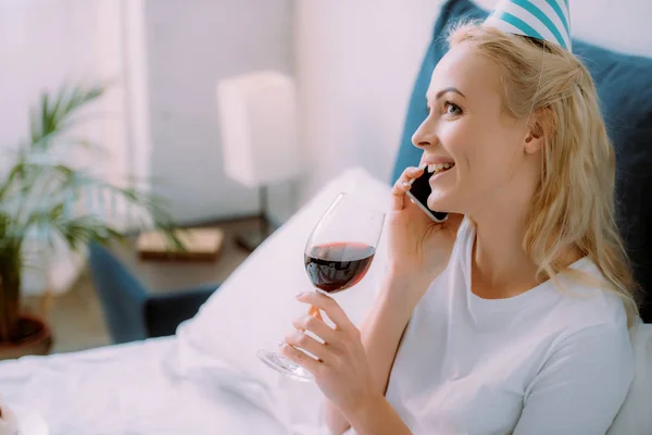 快乐的女人在党的帽子庆祝生日 拿着一杯葡萄酒 在智能手机上在家里的床上交谈 — 图库照片