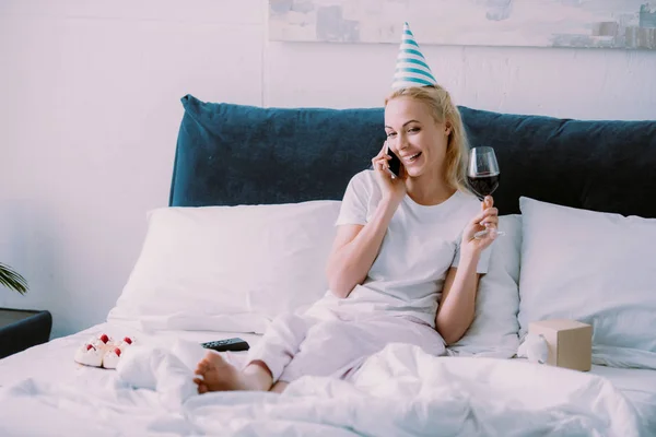 パーティー ハットの誕生日を祝って ワインのグラスを持って 自宅のベッドでのスマート フォンの話で幸せな女 — ストック写真