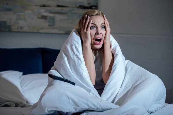 scared woman covered in blanket with hands on head screaming while watching tv in bed at home