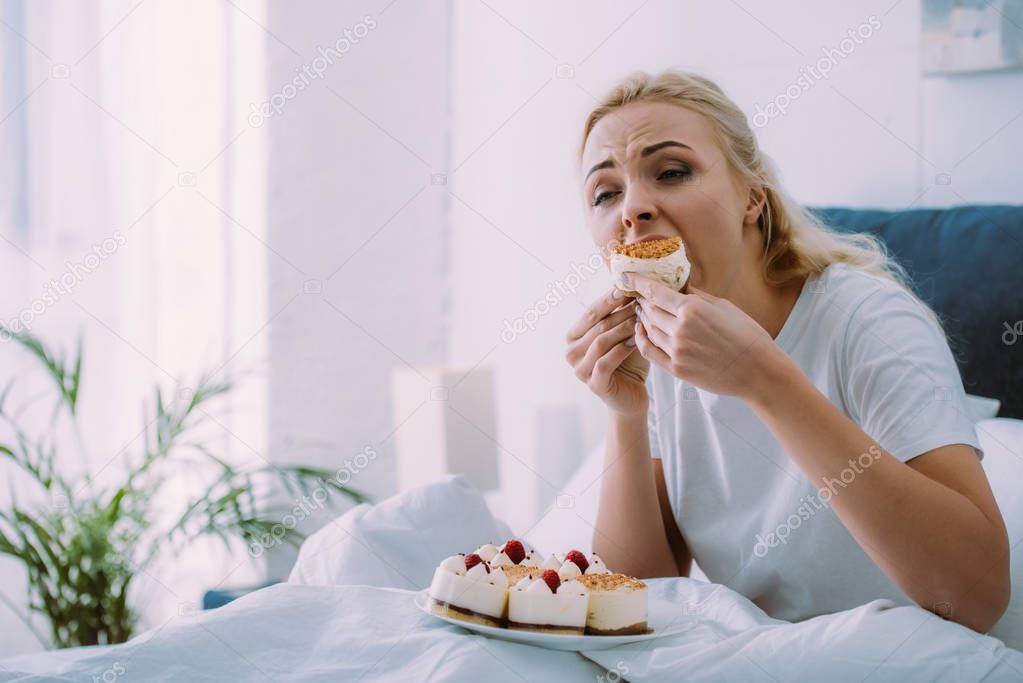 selective focus of sad woman in pajamas eating cake in bed alone