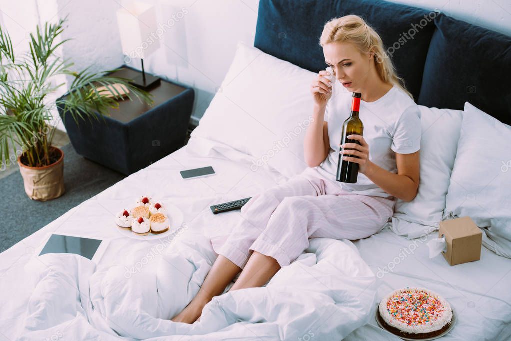 upset woman with bottle of wine wiping tears while celebrating birthday in bed alone