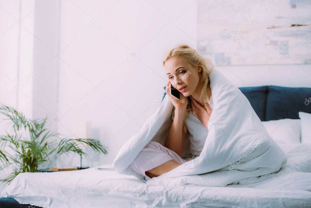 upset woman covered in blanket talking on smartphone in bed at home with copy space