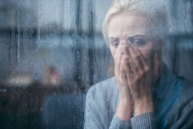 sad adult woman crying and covering face with hands at home through window with raindrops clipart
