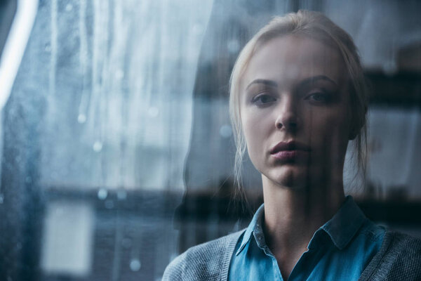 selective focus of sad adult woman at home looking at camera through window with raindrops