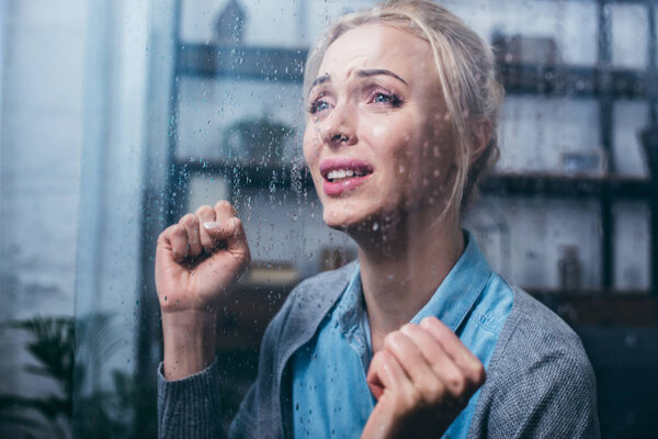 sad adult woman crying with clenched fists at home through window with raindrops