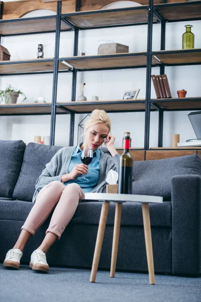 sad woman sitting on couch with glass of red wine and tissue box in living room