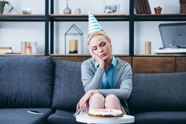 sad woman in party hat propping head with hand while celebrating birthday at home alone