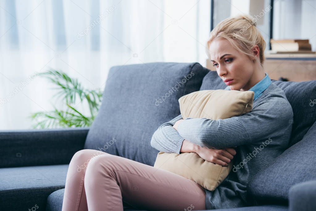 depressed woman sitting on couch and holding pillow at home