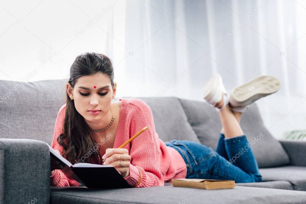 beautiful indian student with bindi writing in notebook and lying on sofa with book