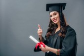 smiling female indian student in academic gown and graduation hat holding diploma and showing thumb up, isolated on grey