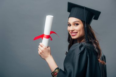 Kız Hint öğrencinin akademik elbisesi ve mezuniyet kap diploma tutan gülümseyerek, gri izole