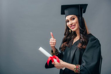 gülümseyen kız Hint öğrencinin diploma ve gösteren başparmak dayanabiliyor, gri izole akademik elbisesi ve mezuniyet şapkalı