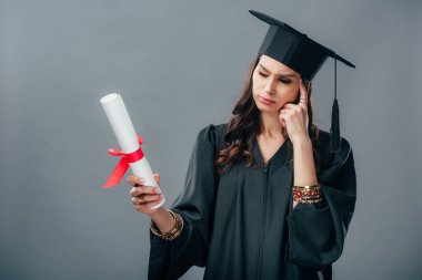 thoughtful indian student in academic gown and graduation hat looking at diploma, isolated on grey clipart