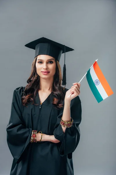 elegant indian student in academic gown and graduation cap holding indian flag, isolated on grey