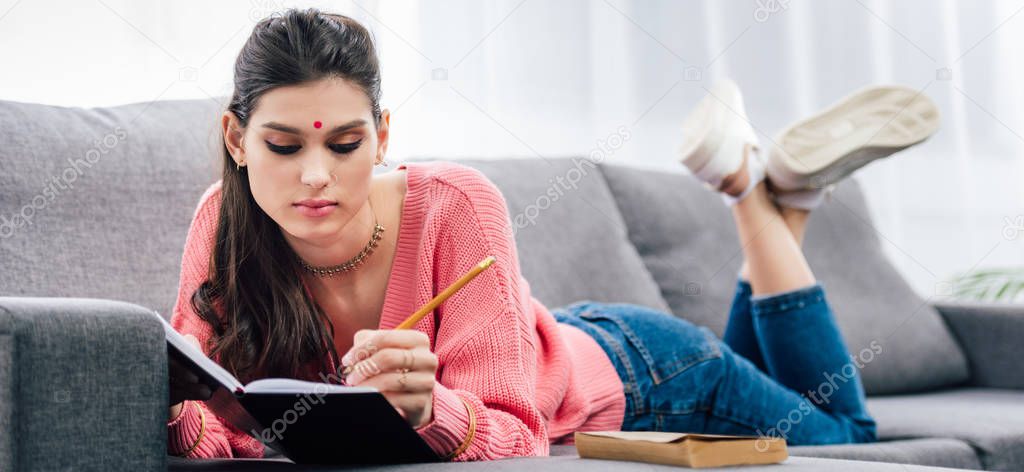 indian student with bindi writing in notebook on sofa with book
