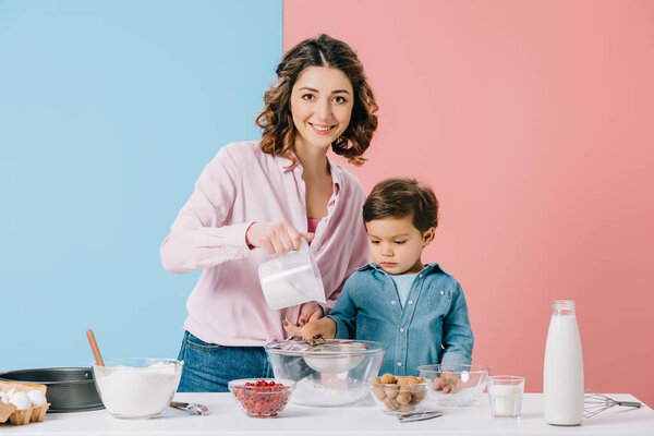 happy mother cooking with little son and looking at camera on bicolor background