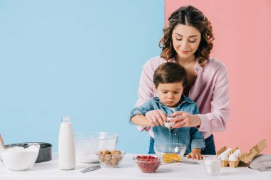konsantre küçük oğluyla birlikte bicolor arka plan üzerinde beyaz mutfak masasında yemek gülümseyen anne 
