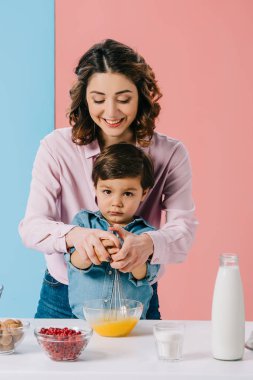 sevimli küçük oğluyla birlikte bicolor arka plan üzerinde pişirme sırasında balon çırpma teli ile yumurta çırpma annesi gülümseyerek