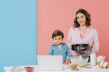 Ürünler, pişirme aletleri ve bicolor arka plan üstünde laptop ile mutfak masasında küçük oğlu ile birlikte yemek yaparken fırın formu tutan anne 
