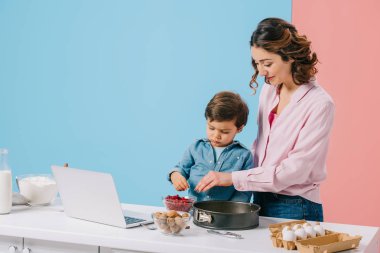 küçük oğluyla birlikte mutfak masasında yemek malzemeler ve bicolor arka plan üstünde laptop ile pişirme sırasında form pişirme için kızılcık ekleyerek annesi gülümseyerek 