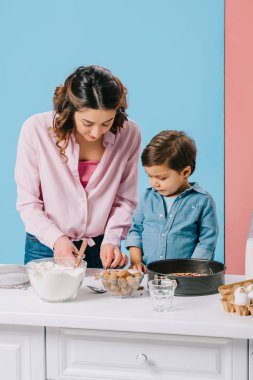 güzel anne ve sevimli küçük oğlu birlikte ürünleri bicolor arka plan üzerinde pişirme ile beyaz mutfak masasında yemek 